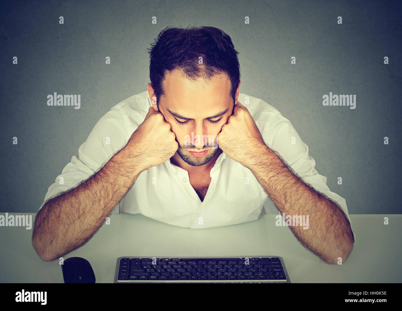 Sad young man sitting at table looking at computer keyboard Stock Photo