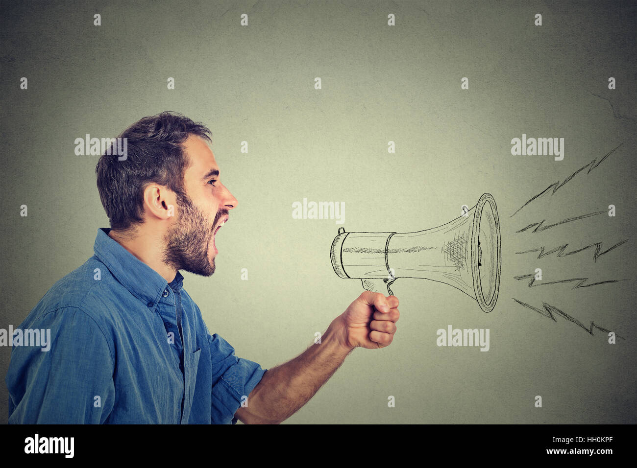 Portrait angry young man holding screaming in megaphone isolated on grey wall background. Negative face expression emotion feeling. Propaganda, breaki Stock Photo