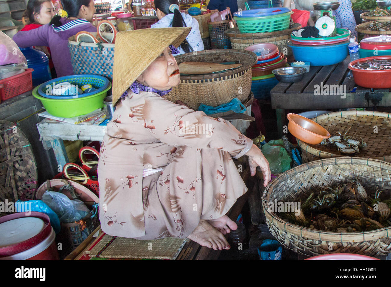 An aged vendor at the fish market wearing the conical hat and silk pyjamas, selling her wares. Stock Photo