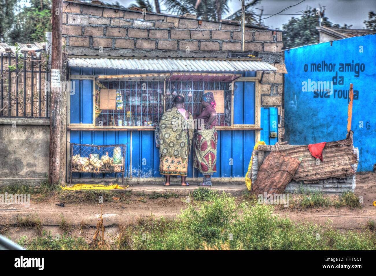 A blue general dealer shop made from tin and brick., with live caged chickens in the front and sarong cladded shoppers. Stock Photo