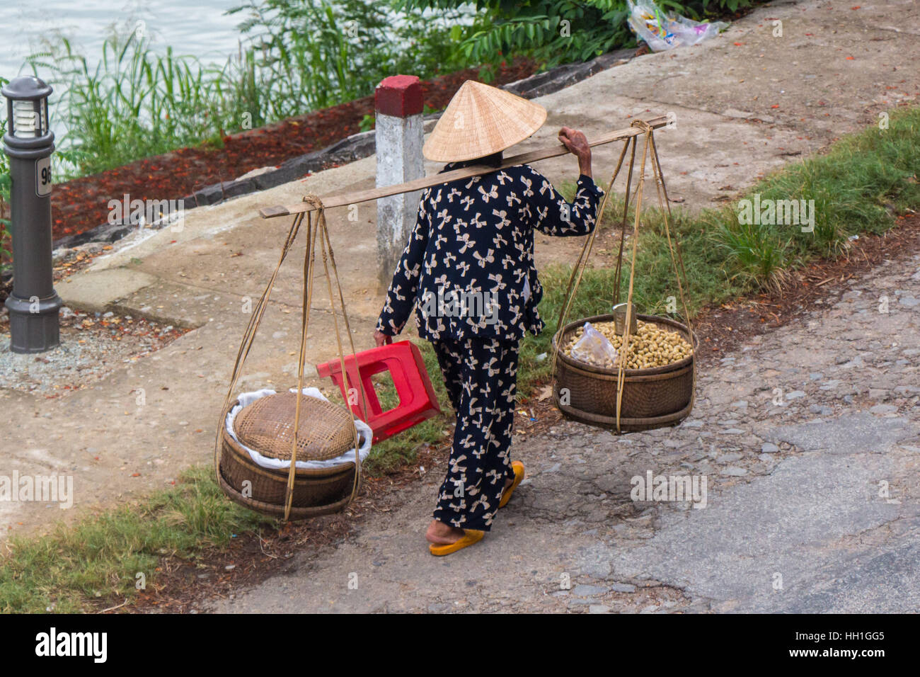 A street vendor carrying her wares in two baskets suspended from a shoulder yoke. Taken along the Perfume River, Hue, Vietnam. Stock Photo