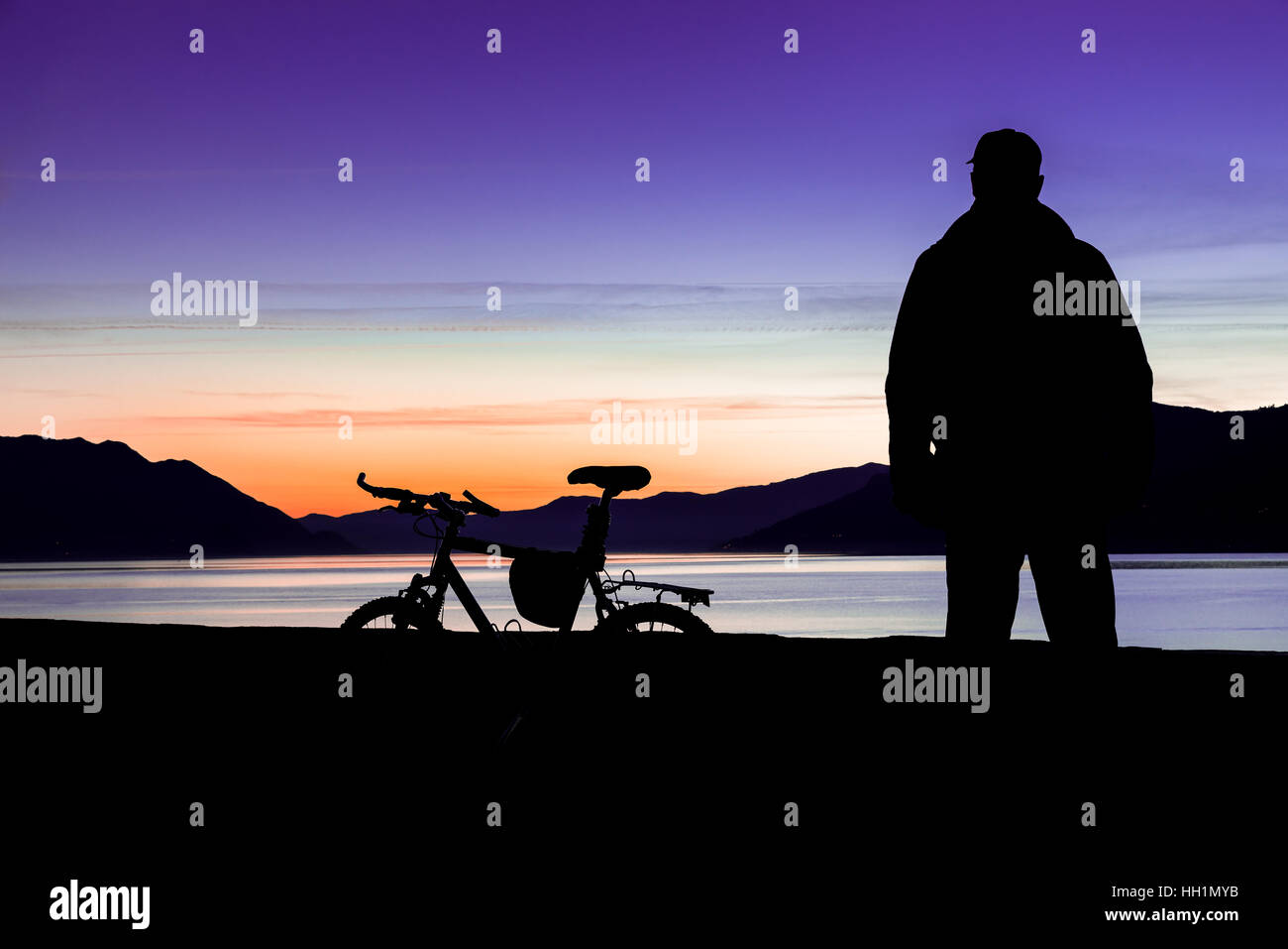 Silhouette of a mountain bike and of a cyclist who is watching the sunset over the lake. Focus on mountain bike Stock Photo