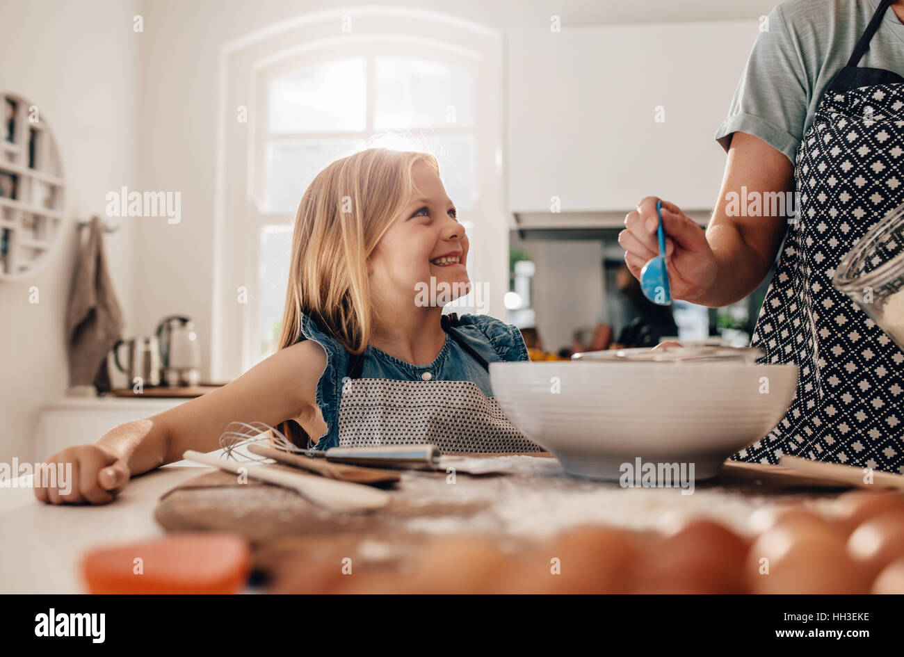 Smiling young girl standing by her mother in kitchen cooking food. Mother and happy daughter baking in kitchen. Stock Photo