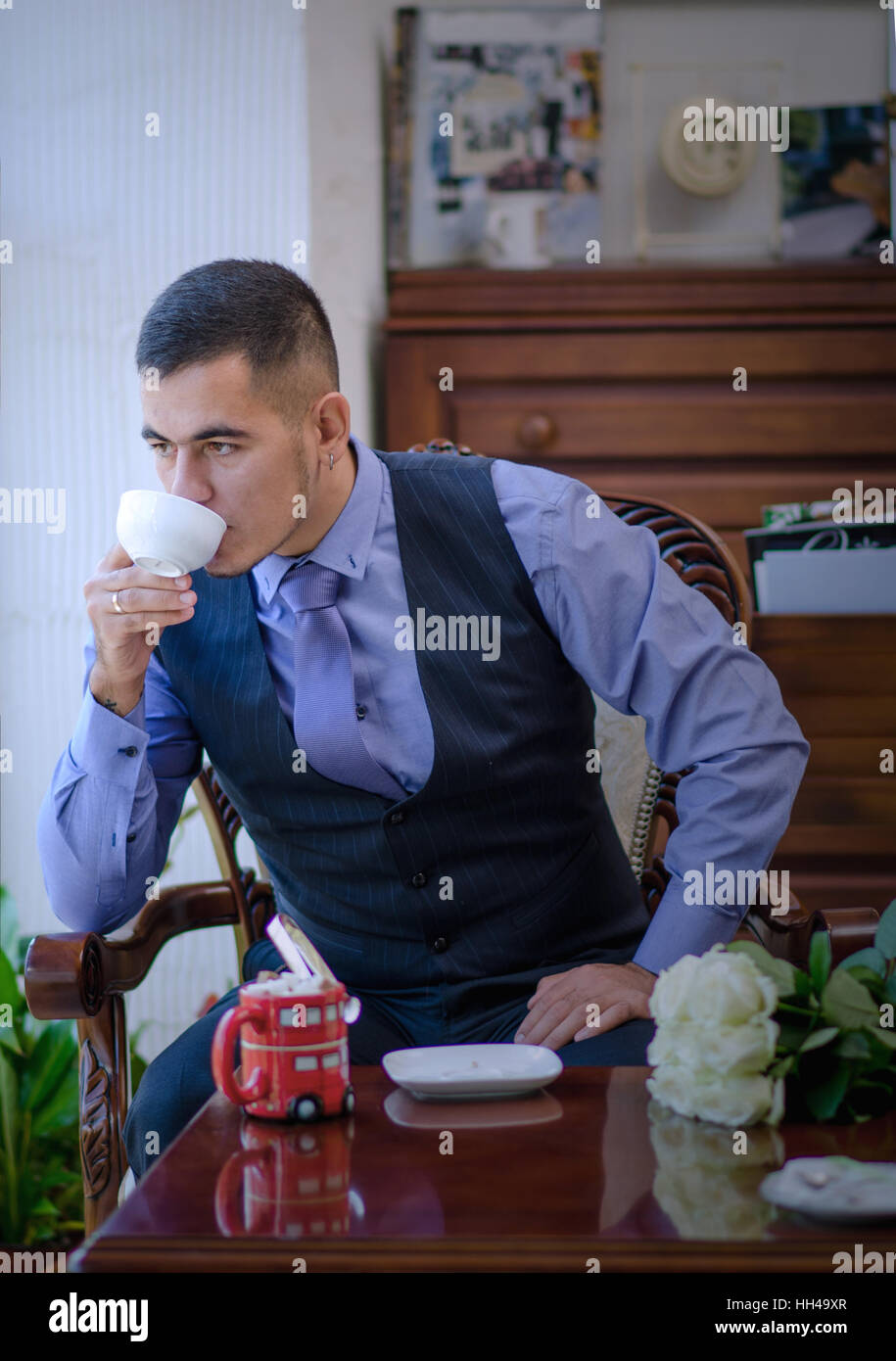 The serious young man in a vest, a tie and with a beard sits on a chair at a window and has tea or coffee Stock Photo