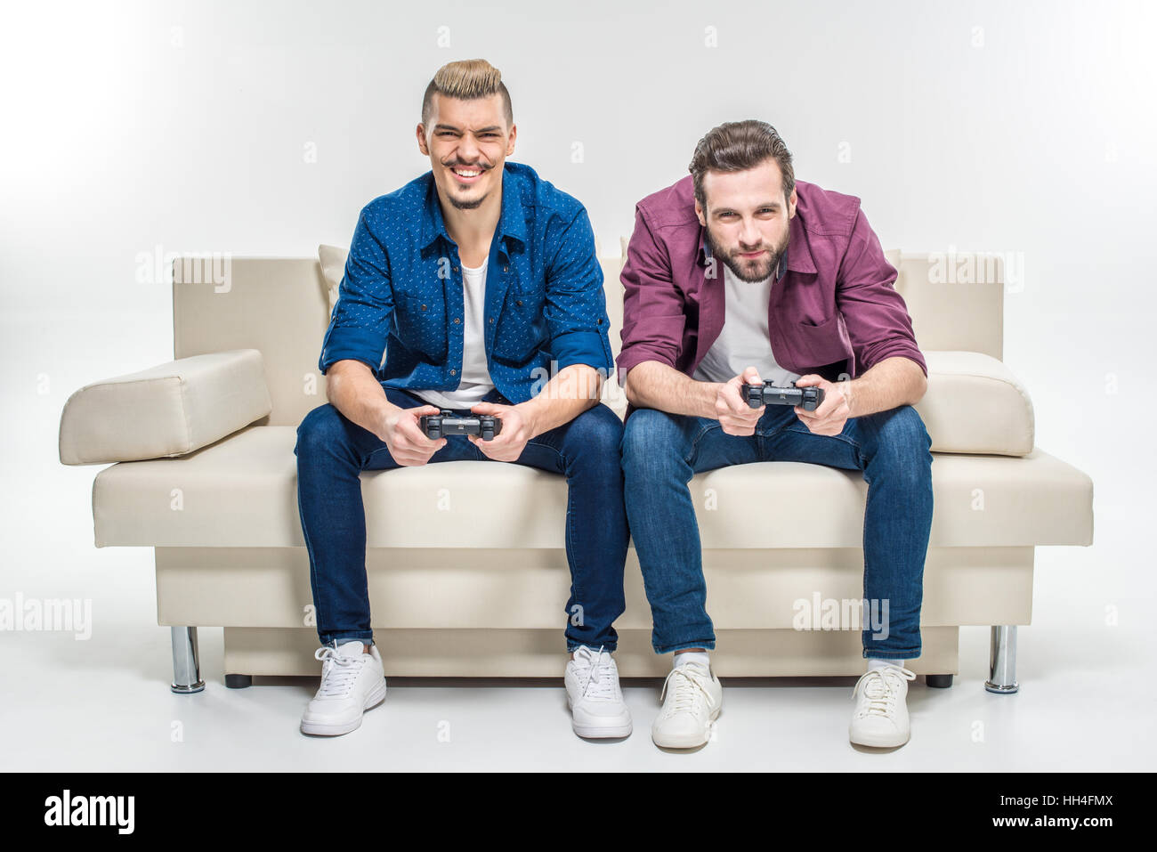 Concentrated male friends sitting on couch and playing with joysticks Stock Photo