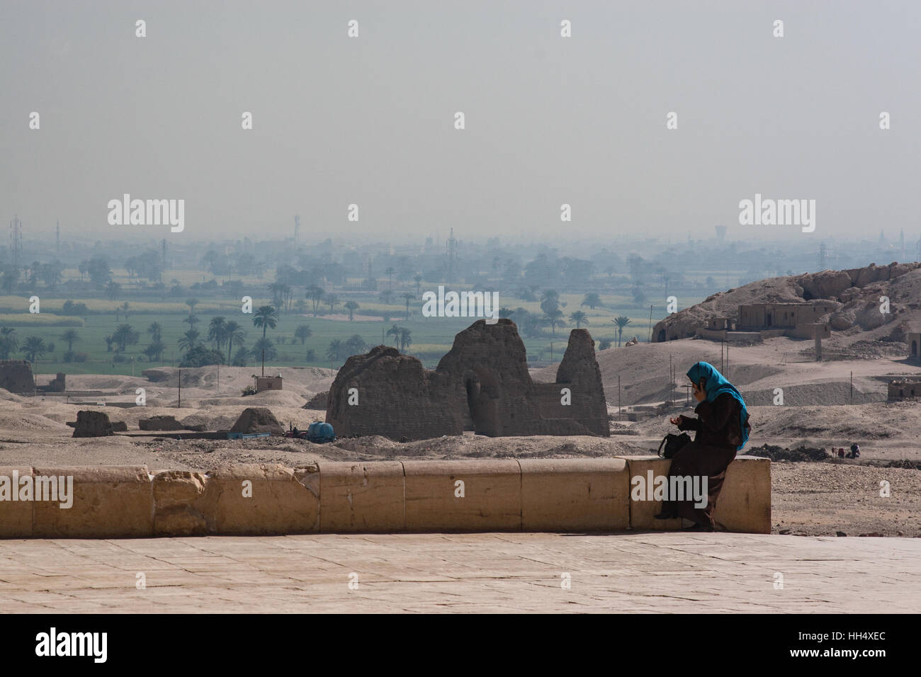 Woman wearing hajib perched on a low stone wall, using a cellphone, outside of the Mortuary Temple of Hatshepsut in Egypt Stock Photo