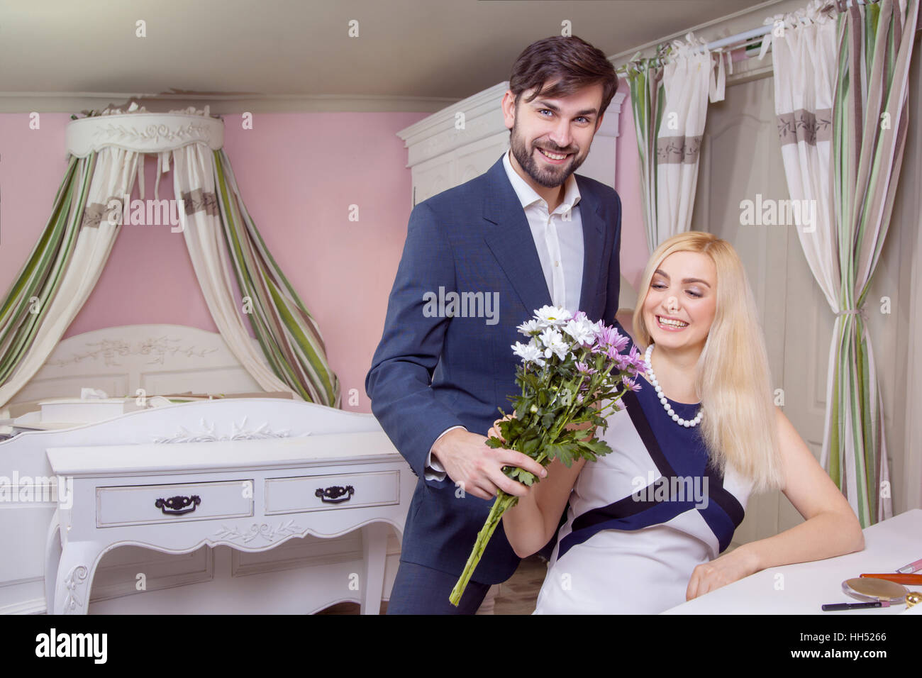 Young handsome man surprising her beautiful girlfriend and gives her bouquet of purple white flowers. Stock Photo