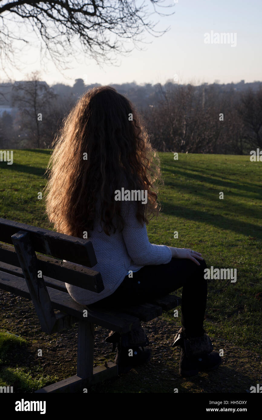 Pensive young woman sitting alone on a bench looking into the distance, in a quiet location with a sky and trees. Stock Photo