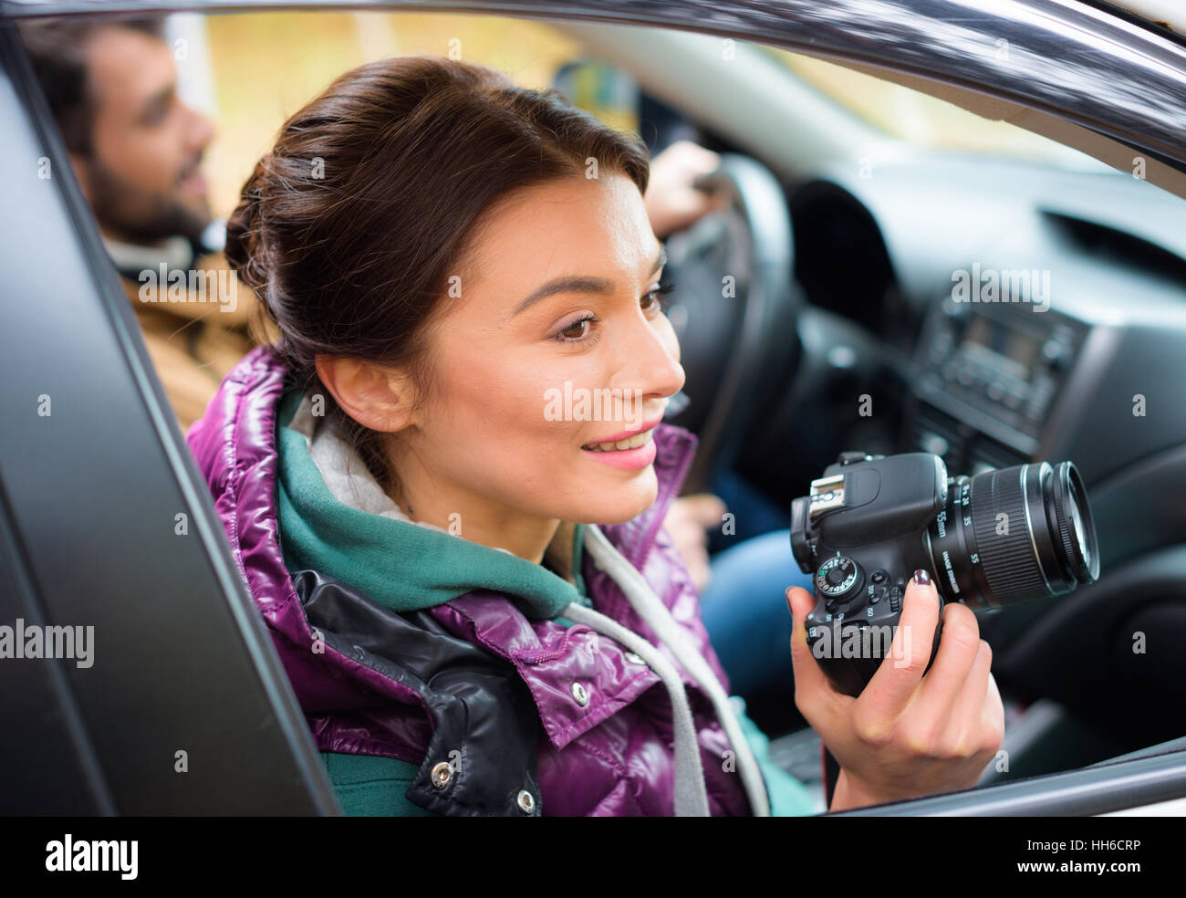 Smiling brunette woman holding photo camera and looking in car window with blurred male driver on background Stock Photo