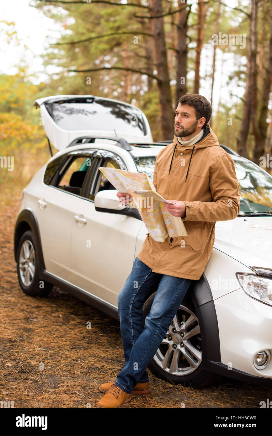 Young bearded man holding map and leaning on white car in autumn forest Stock Photo
