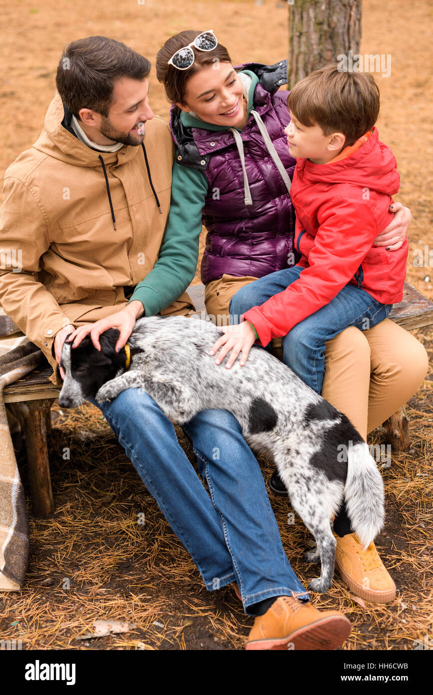 Happy family sitting on wooden bench and stroking dog in autumn forest Stock Photo