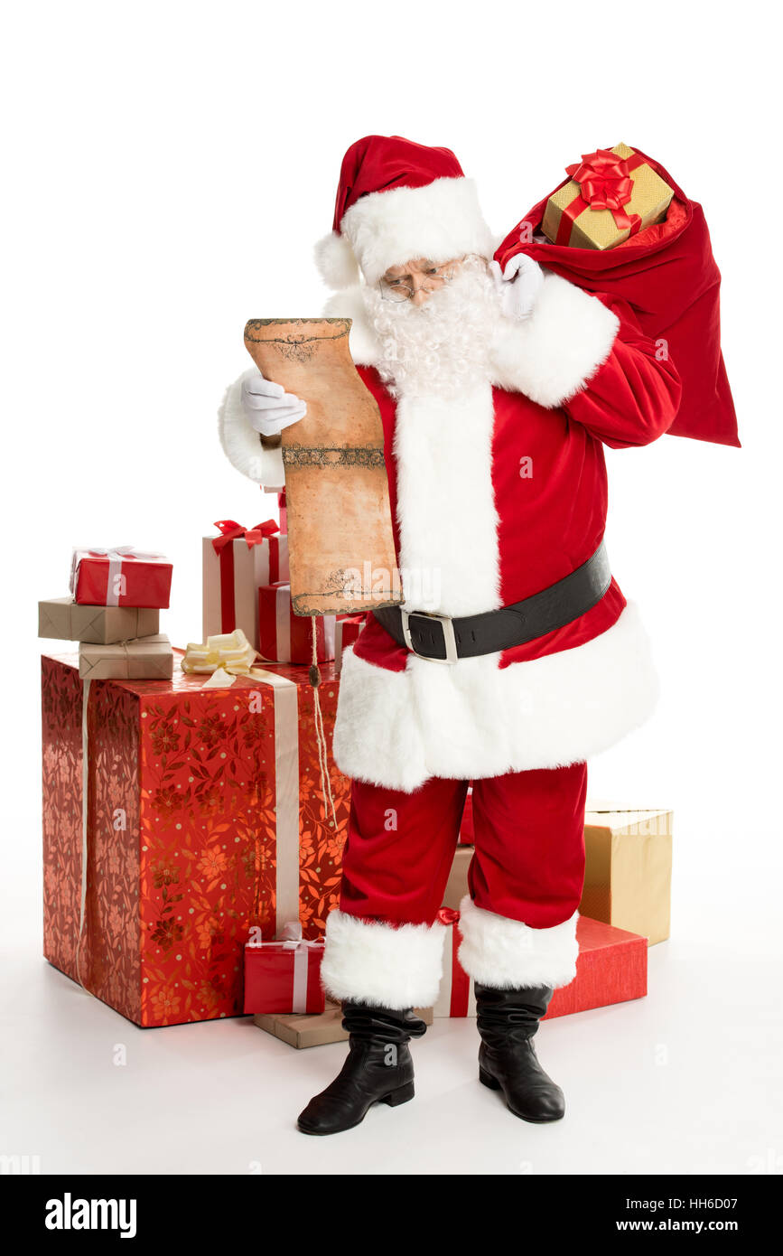 Happy Santa Claus standing with a pile of Christmas gifts and a big sack on back reading wishlist Stock Photo