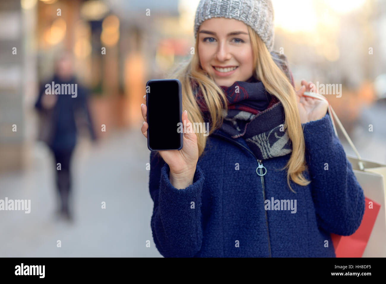 Pretty young woman holding up her mobile phone with a blank display as she stands on an urban street in winter warmly dressed in a knitted woollen sca Stock Photo