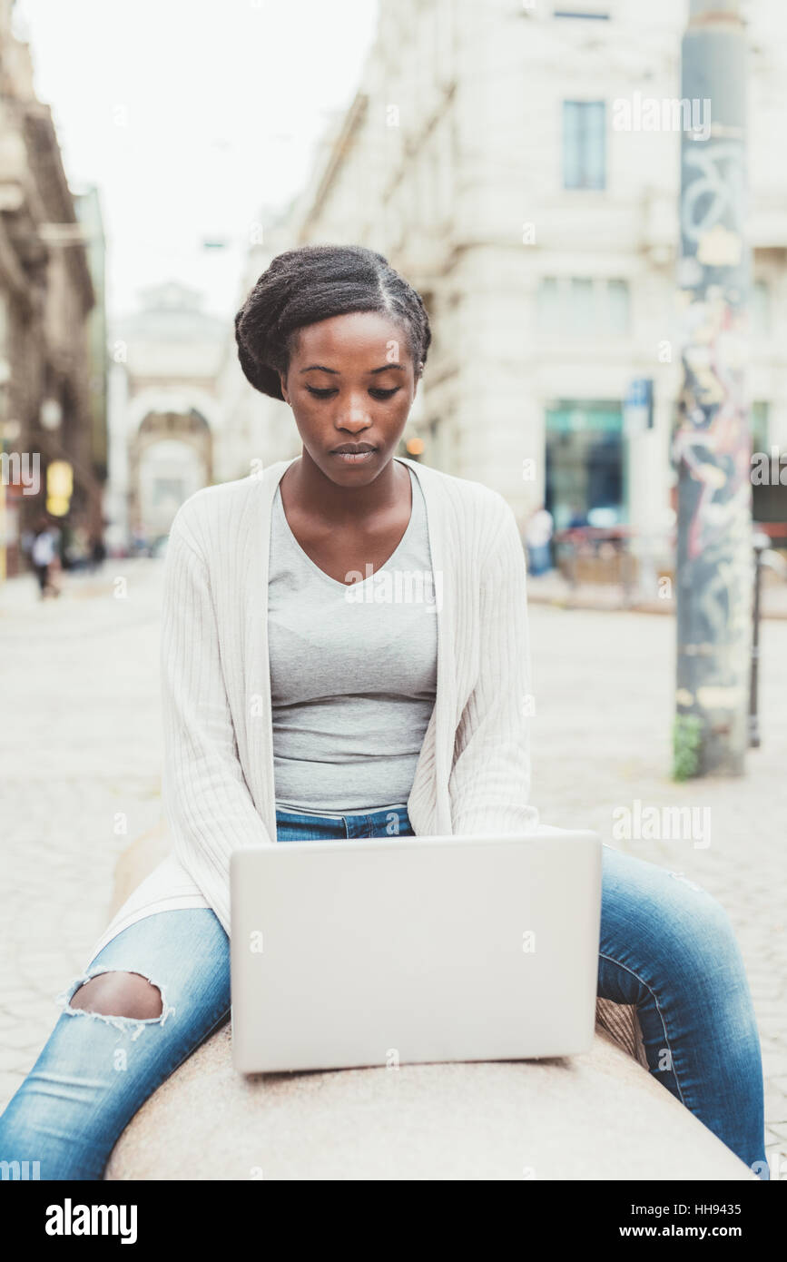 Young black woman using computer outdoor in the city sitting on a bench ...
