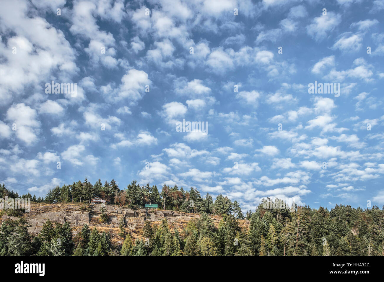 A bright blue sky with many small clouds, above a forested rocky ridge with two homes on its edge. Stock Photo