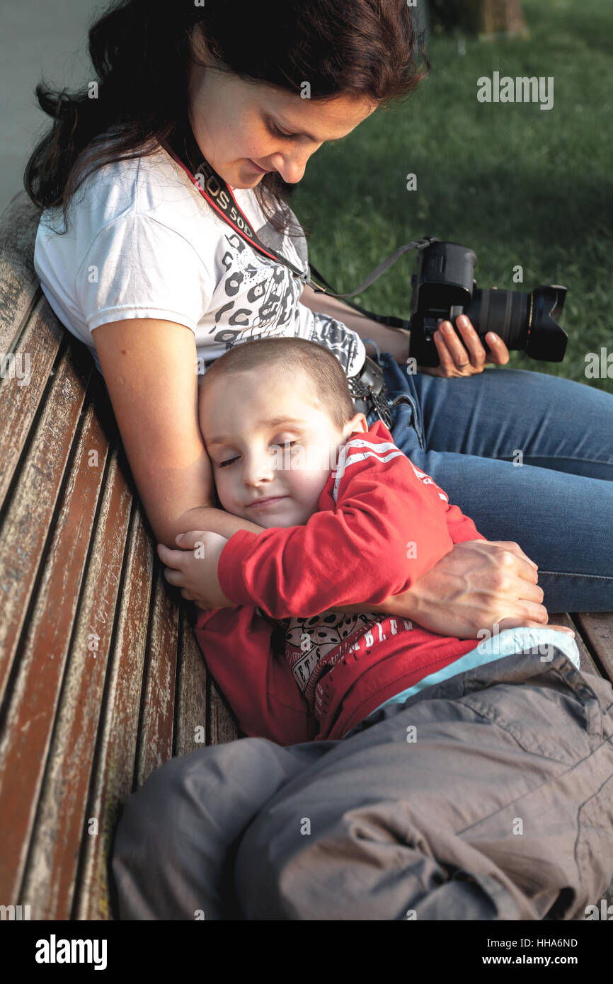 Young kid boy laying and sleeping in mother's lap on a bench Stock Photo