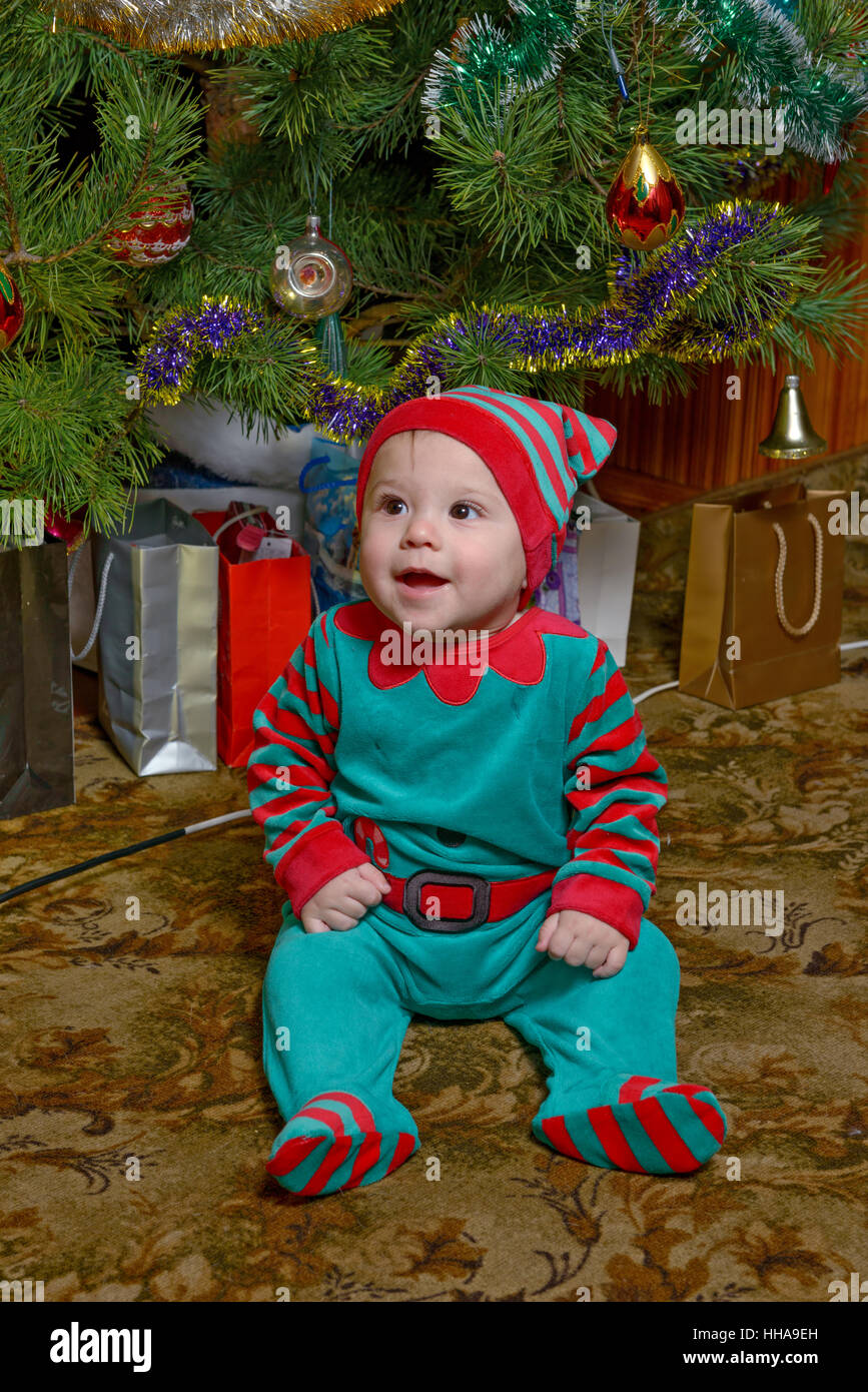 Smiling little baby boy in red-green suit is sitting on floor napless carpet covering near Christmas tree. Stock Photo