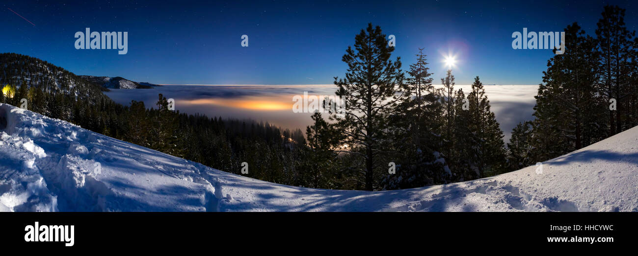 Cold Winter Snowy landscape at night with cloud inversion covering city lights that glow underneath the cloud cover.  Lit with moonlight and the sky h Stock Photo
