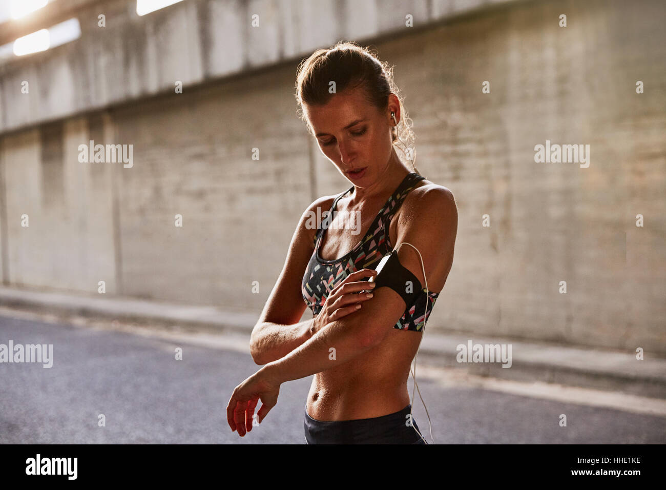 Fit female runner in sports bra using mp3 player arm band and headphones on urban street Stock Photo