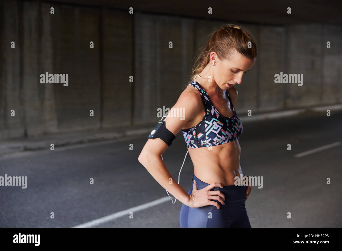 Fit female runner in sports bra with mp3 player armband and headphones resting in urban tunnel Stock Photo