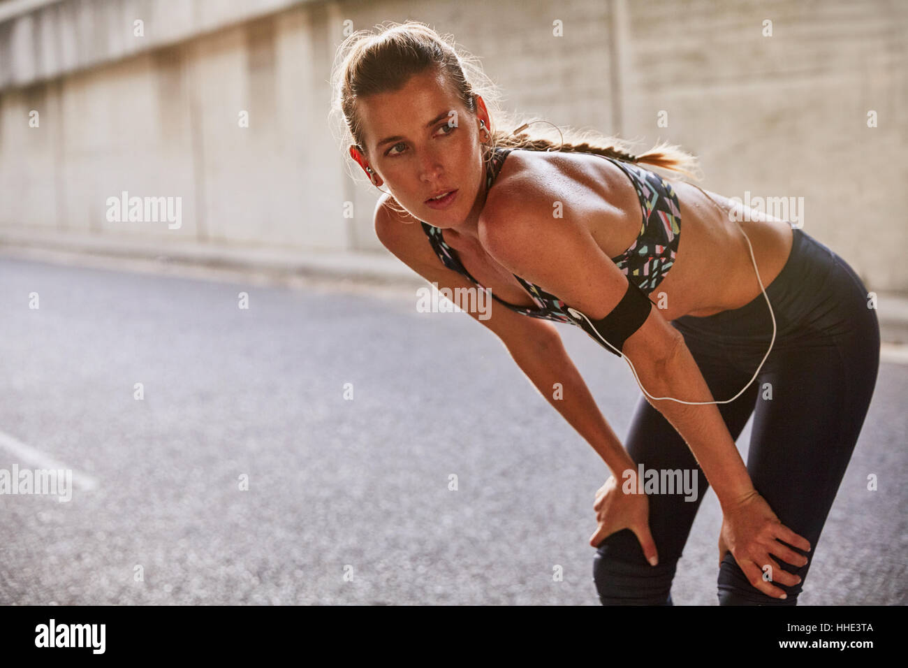 Tired fit female runner in sports bra with mp3 player armband resting on urban street Stock Photo