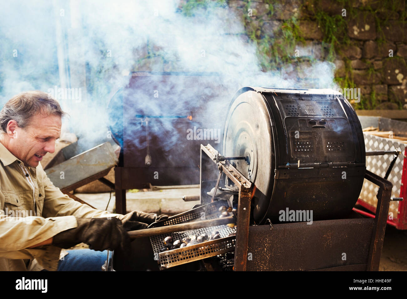 A man roasting chestnuts over hot coals in a large drum, raking out the