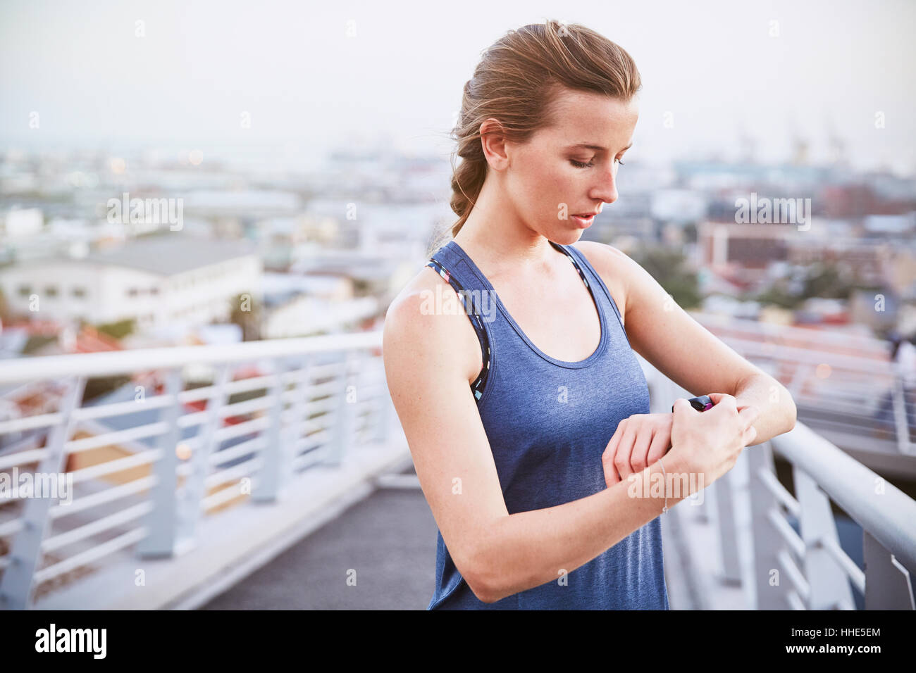 Female runner checking smart watch fitness tracker on urban footbridge Stock Photo