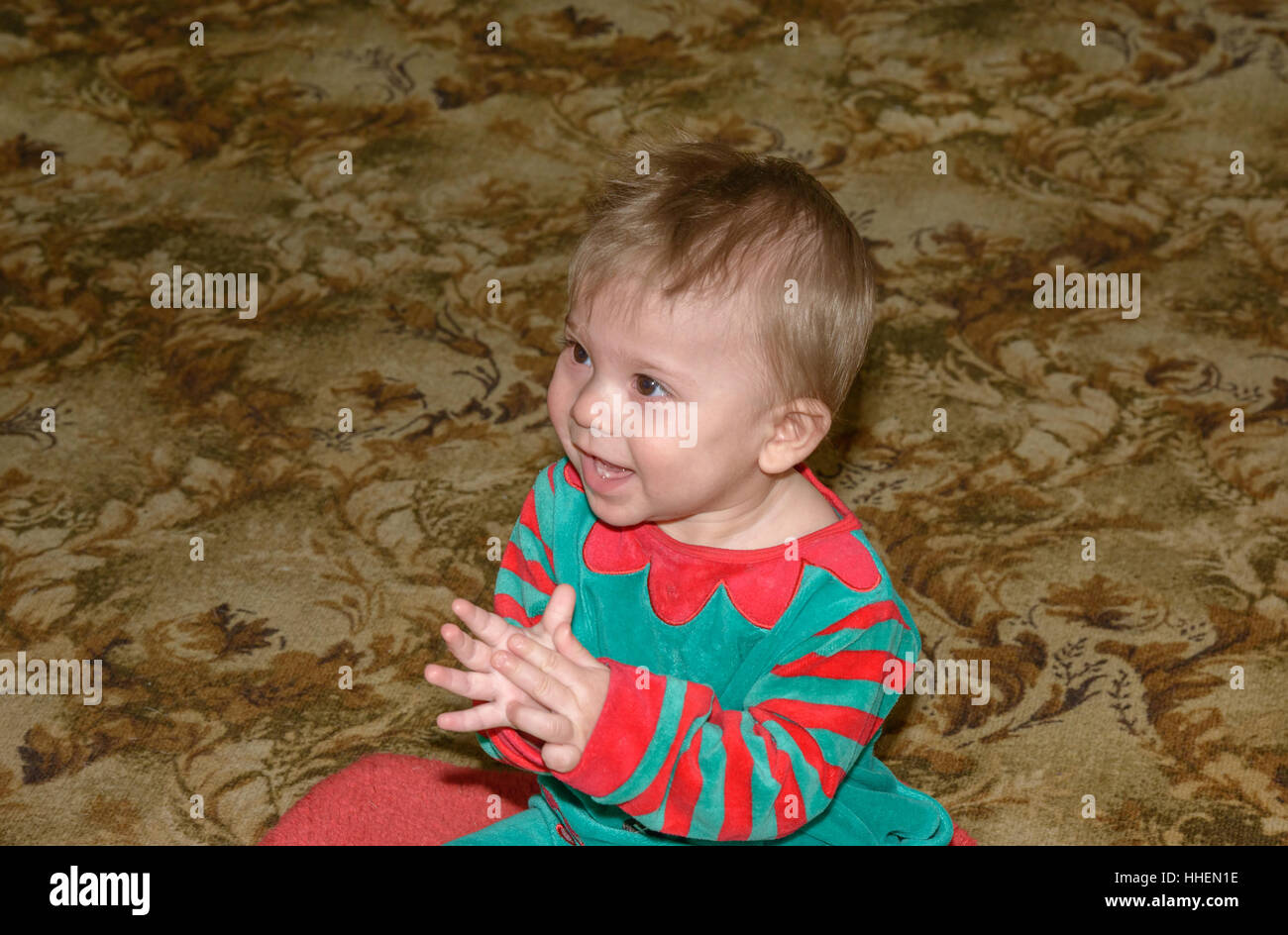 Little baby boy in red-green suit is learning to play patty-cake sitting on floor napless carpet covering. Stock Photo