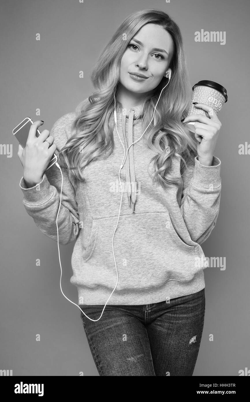 Portrait of a cute positive blonde woman wearing gray hoodie in headphones listening music with phone and coffee on bright background in studio Stock Photo