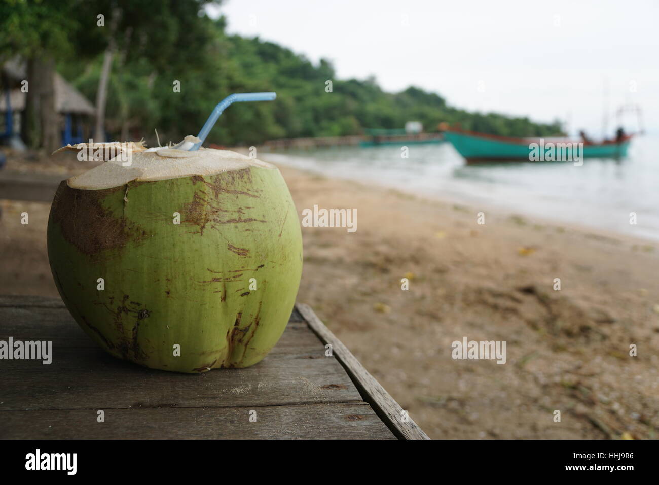 Coconut with Straw on a Bench at a Tropical Beach with Boat in the Background Stock Photo