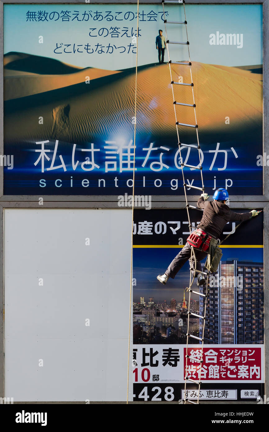 A workman puts up posters on an advertising billboard below an existing billboard for the Church of Scientology,. Shibuya, Tokyo, Japan. Stock Photo