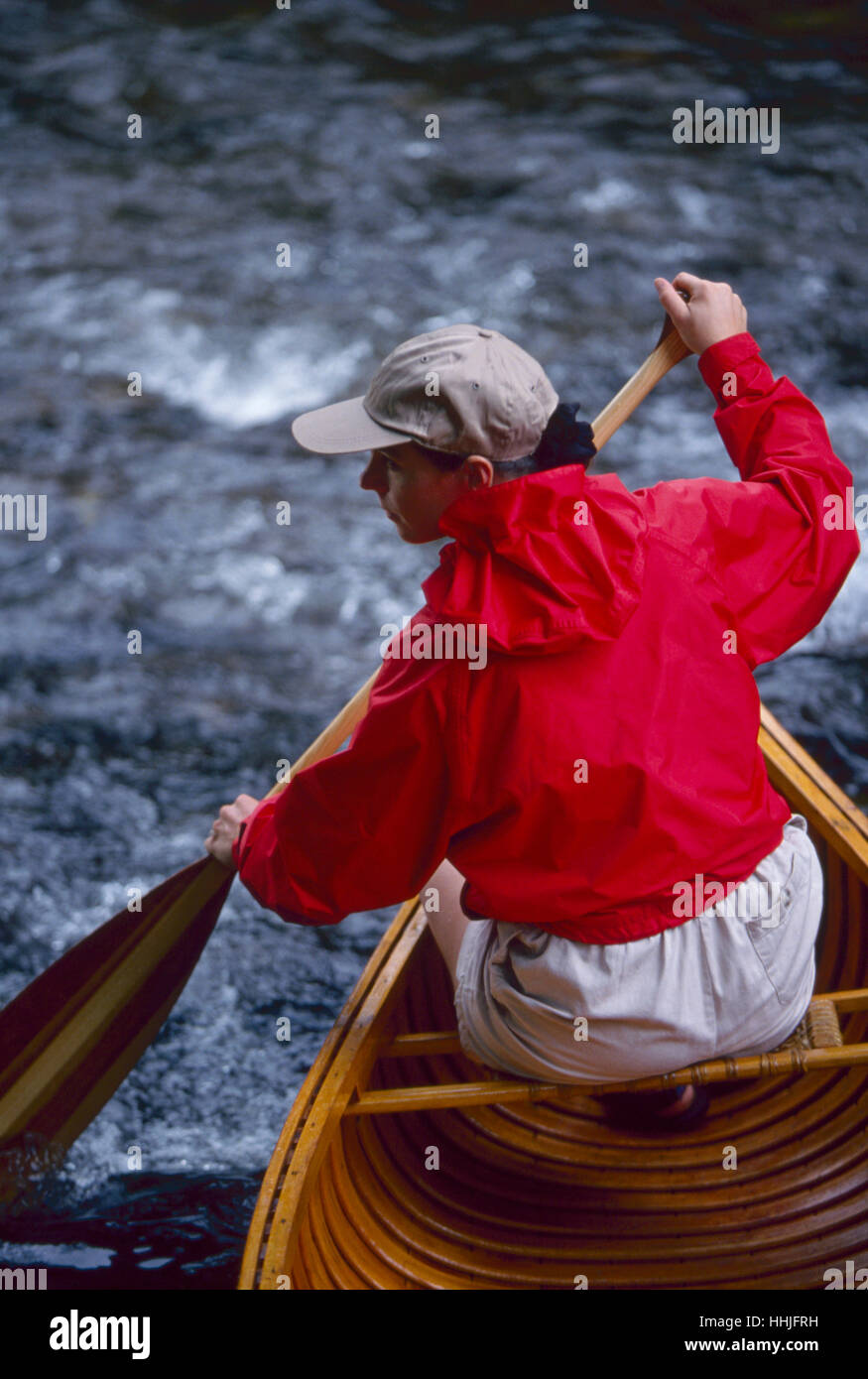 A young woman in her 20's wearing a bright red jacket paddles her vintage wood canoe in a  whitewater stream Stock Photo