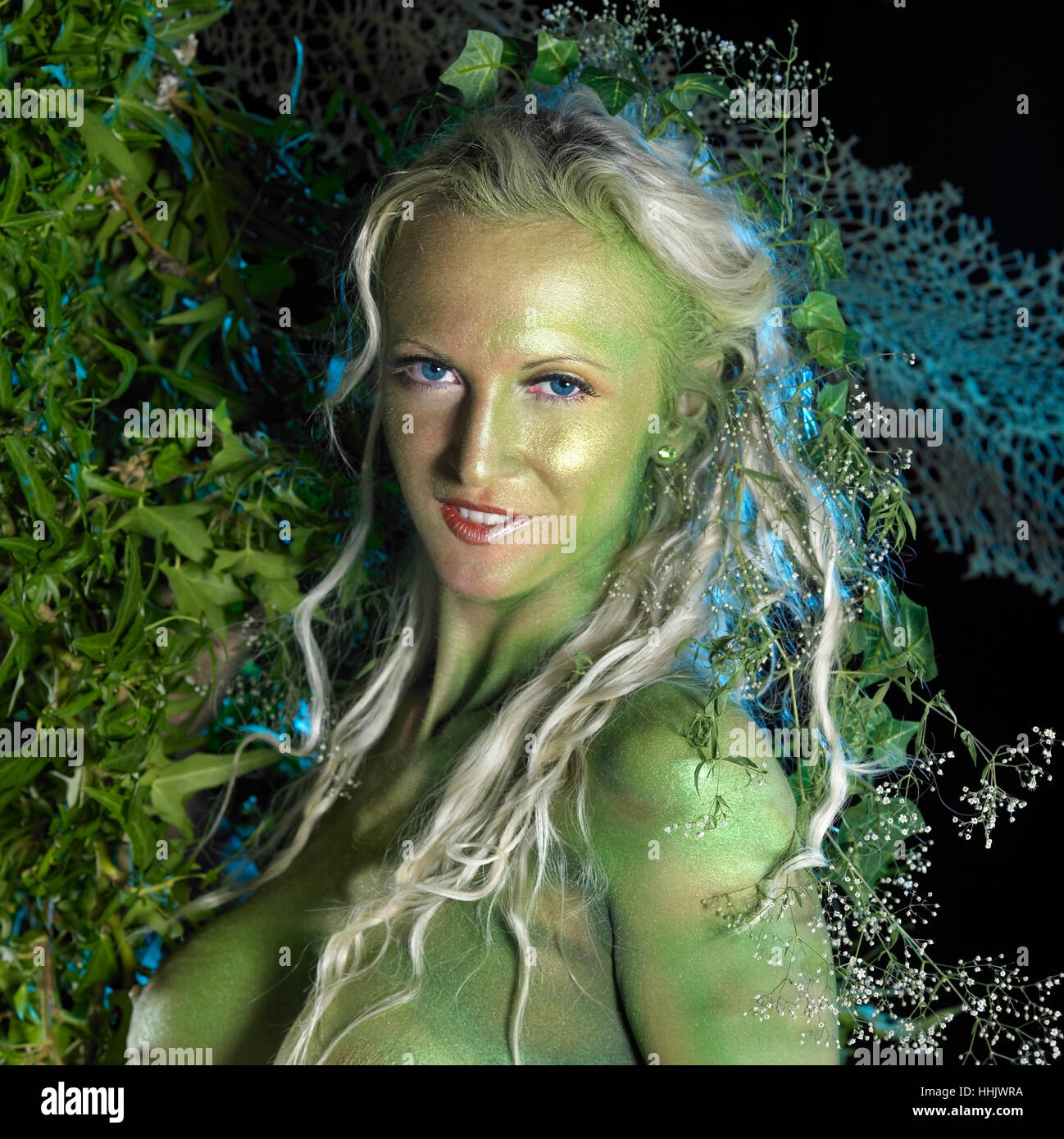 bodypainted woman in front of dark back, partly hidden by green vegetation Stock Photo