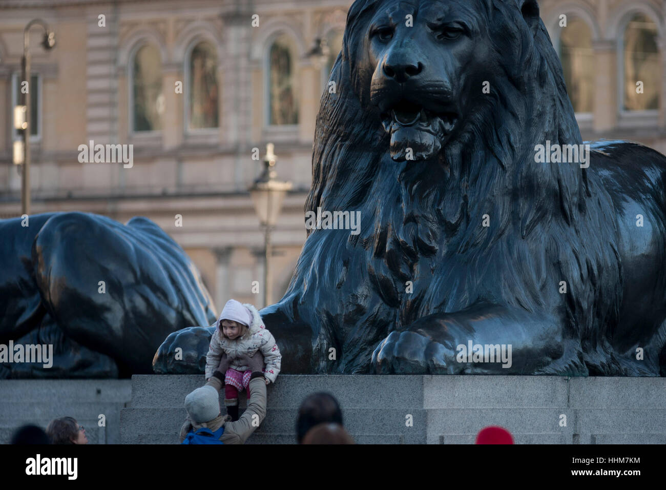 A young girl sits beneath one of the four enormous lion statues at the base of Nelson's column, on 17th January 2017, in Trafalgar Square, London England. The column dedicated to the heroic naval Admiral Lord Nelson is guarded by the four monumental bronze lions sculpted by Sir Edwin Landseer. In recent years there have been numerous falls from the lions resulting in serious injury including the necessity of the air ambulance. Stock Photo