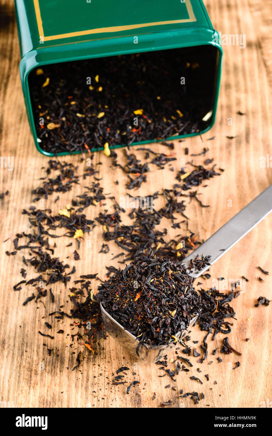 Flavored black tea in overfull metal scoop on wooden board. Open green tea canister in background. Stock Photo