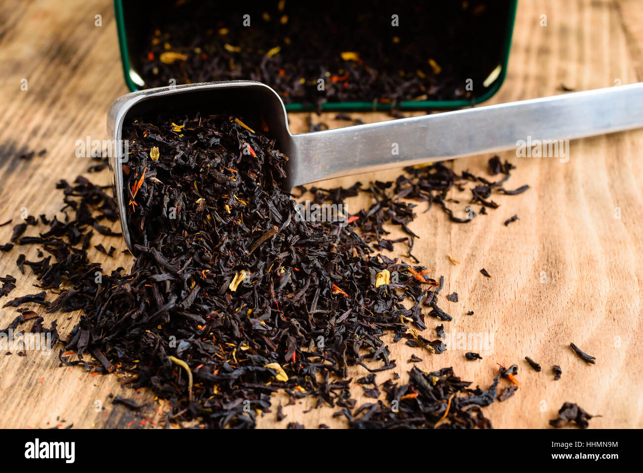 Flavored black tea spilling out of metal scoop on wooden board. Open tea canister in background. Stock Photo