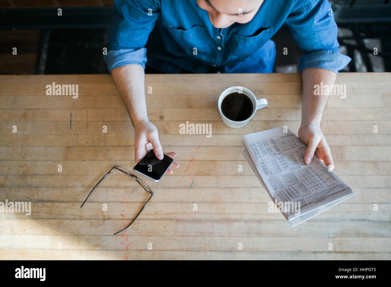 Young man reading paper and phone, drinking coffee, sitting at a wooden table in hipster cafe and looking down, glasses on table Stock Photo