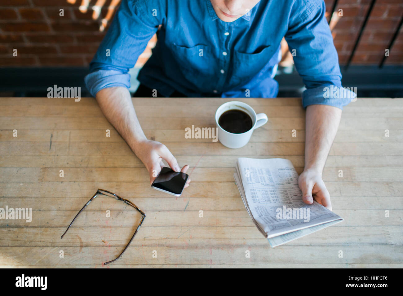 Young man reading paper and phone, drinking coffee, sitting at a wooden table in hipster cafe and looking down, glasses on table Stock Photo