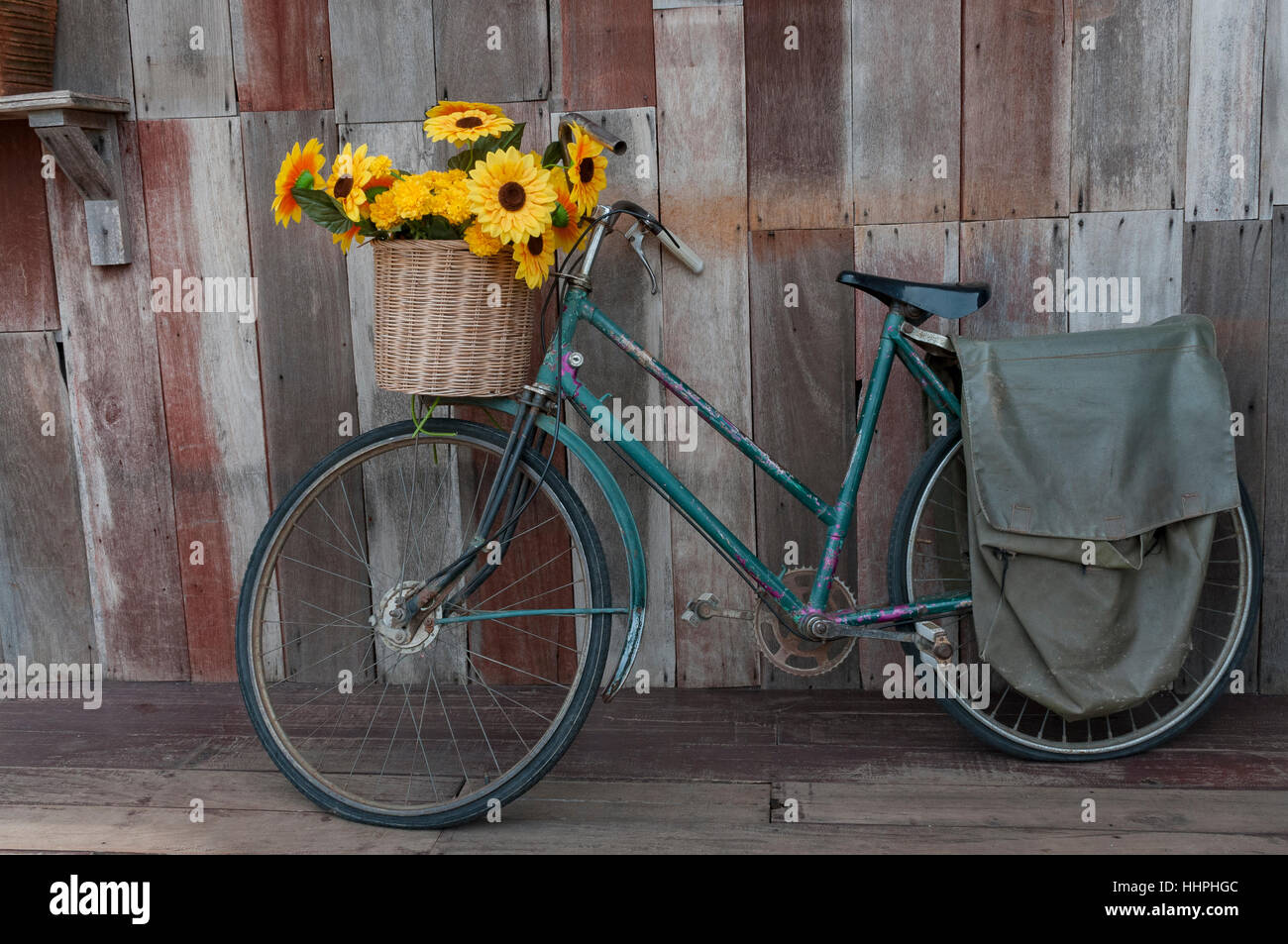An old pedal bicycle, with a basket full of yellow flowers, leaning against the wooden wall in Nong Khai, Northeast Thailand. Stock Photo