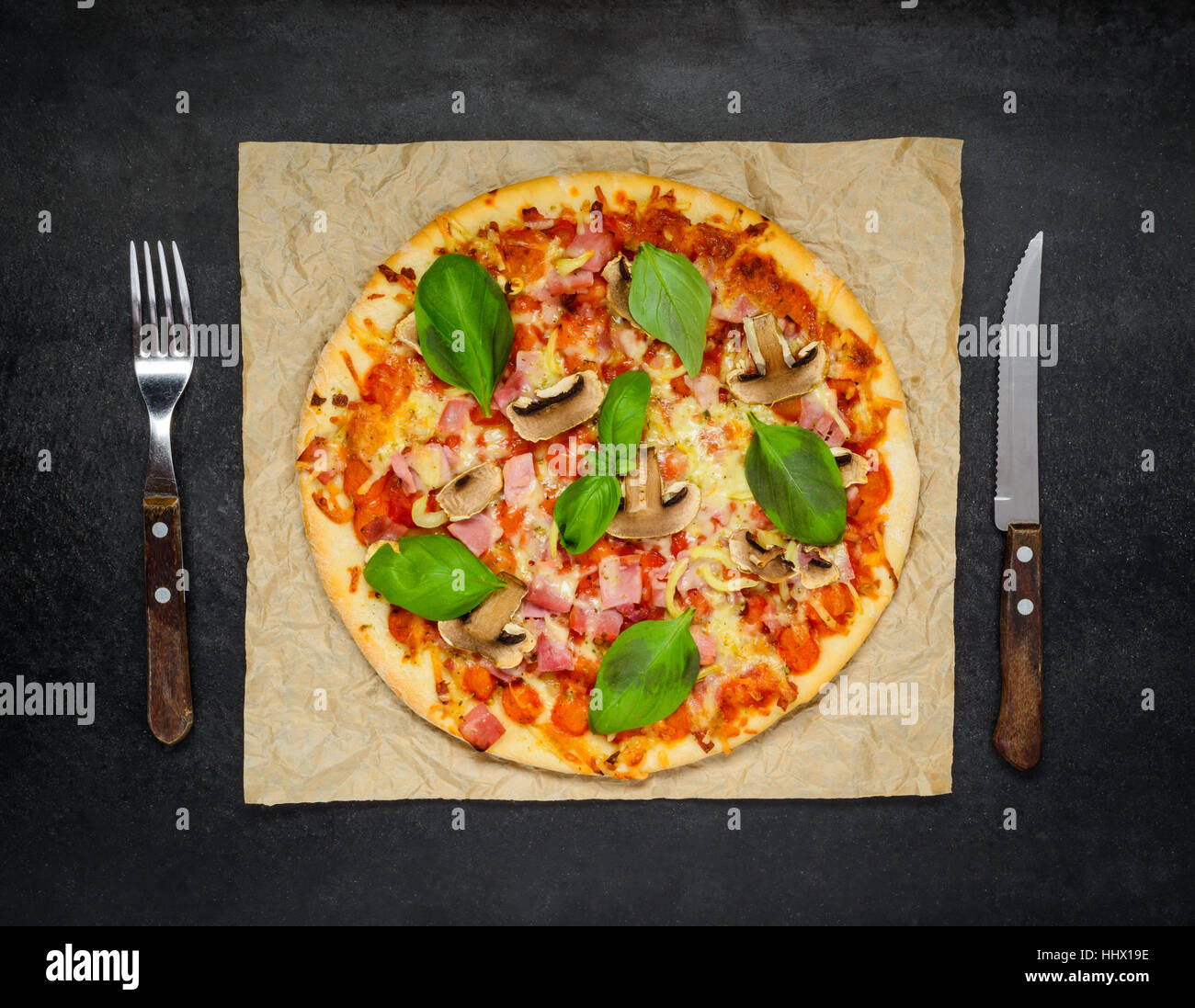 Fresh Baked Pizza with mushrooms, cheese and basil and Fork and Knife on a Dark Stone Table. Stock Photo