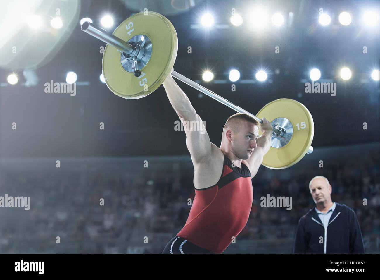 Coach watching male weightlifter squatting barbell overhead in arena Stock Photo