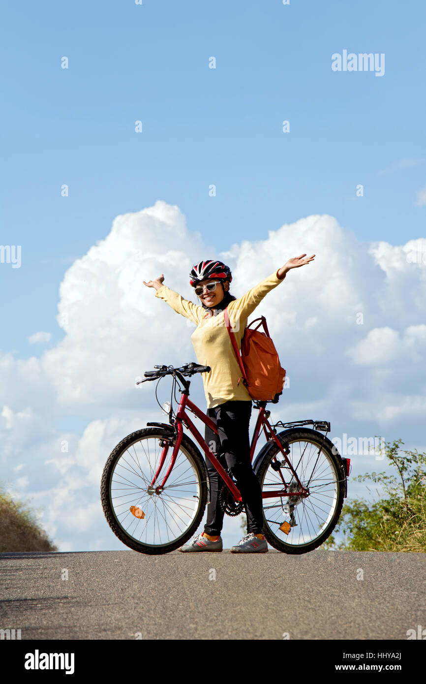 Woman cyclist standing with a bicycle on the road horizon and hands lifted up to heaven. Stock Photo