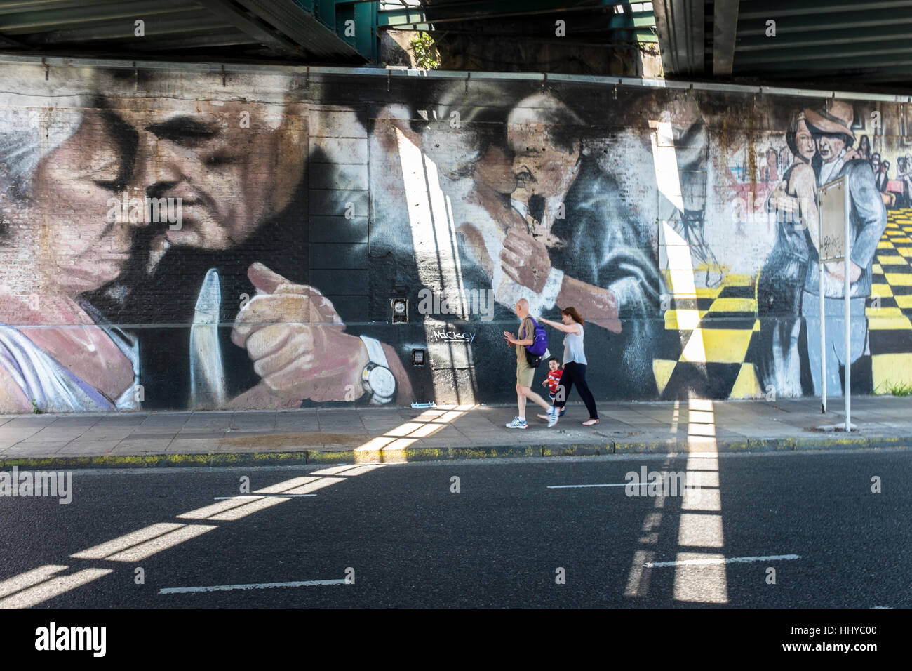 Pedestrians walk under a railway bridge where painted walls depict three couples dancing Tango in Buenos Aires. Stock Photo