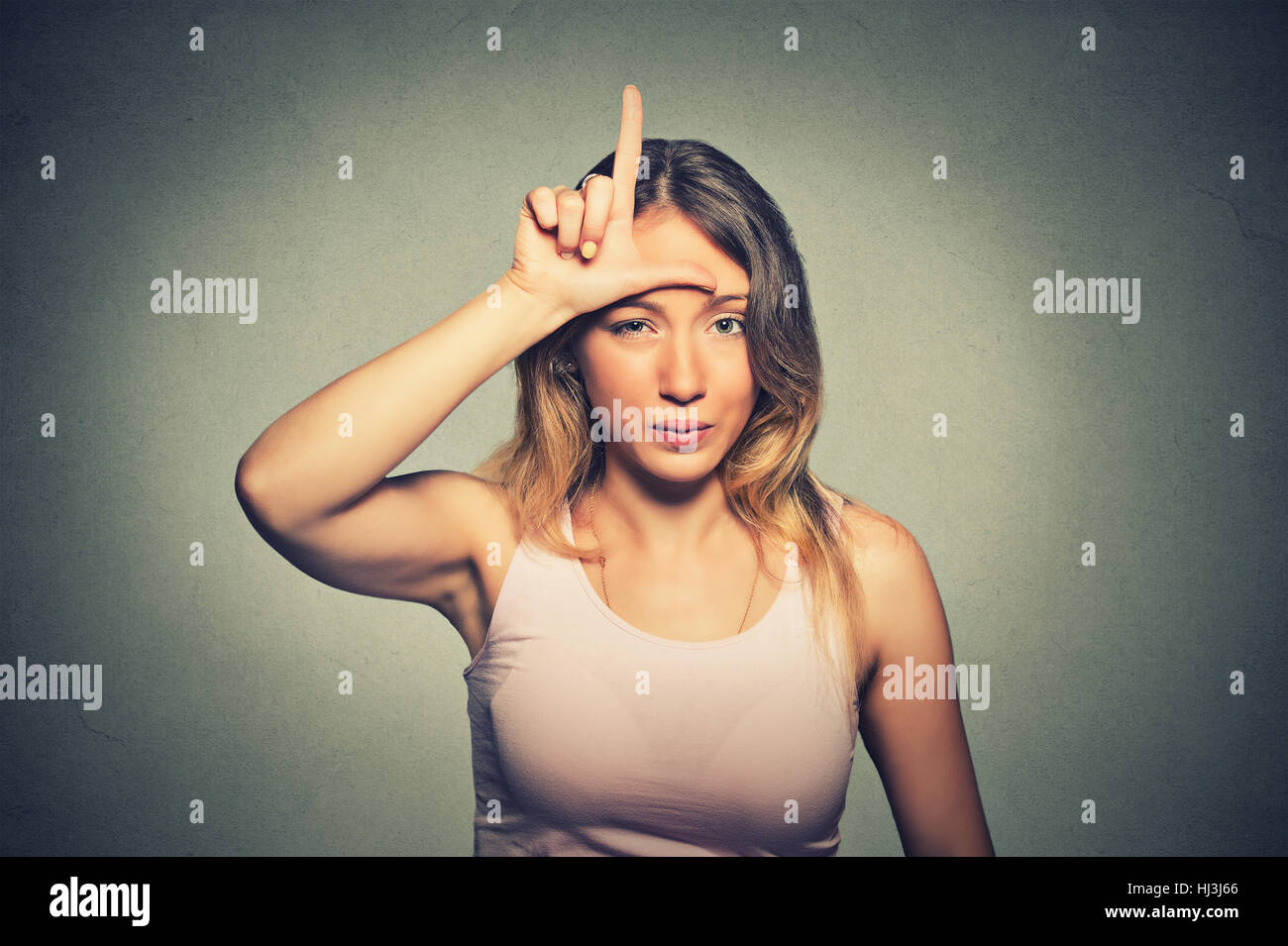 Closeup portrait young unhappy woman giving loser sign on forehead, looking at you, disgust on face isolated on gray wall background. Negative human e Stock Photo