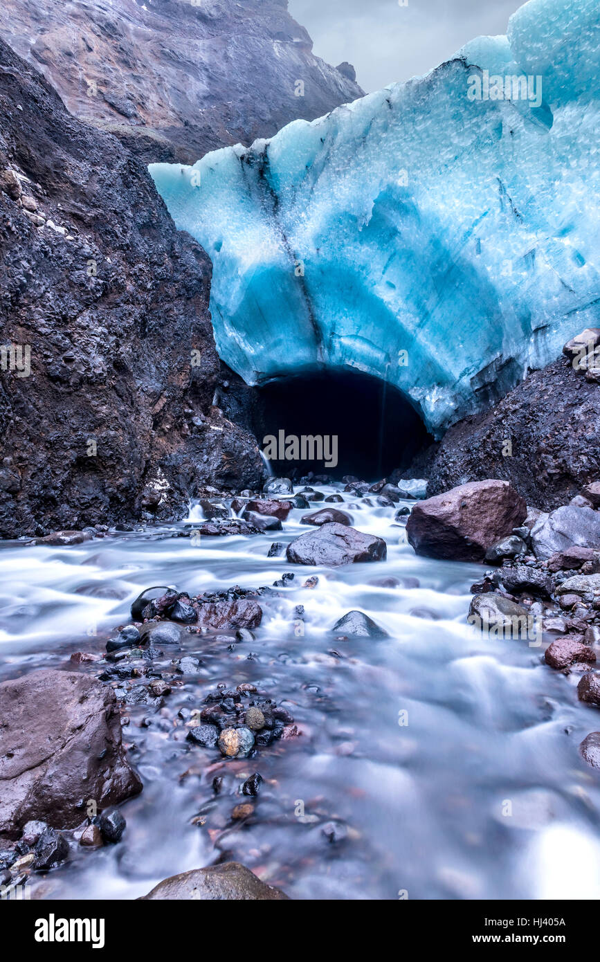 An ice cave in Iceland formed below a glacier is melting and forming a river lowing through the mouth of the cave. Stock Photo
