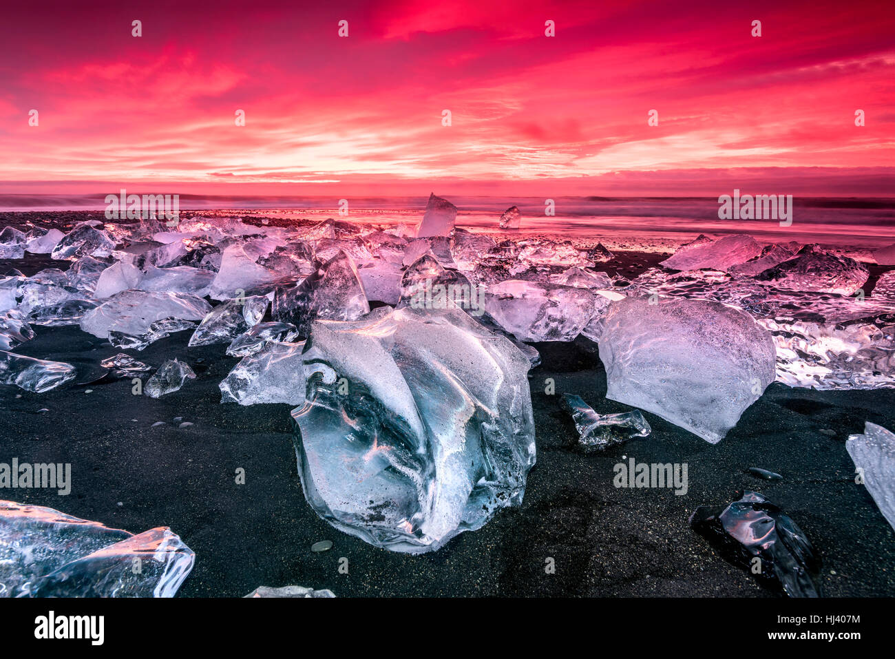 An iceberg along the shore of Jokulsarlon glacial lagoon during a vibrant red sunrise rests motionless as it is framed by cold ocean water. Stock Photo