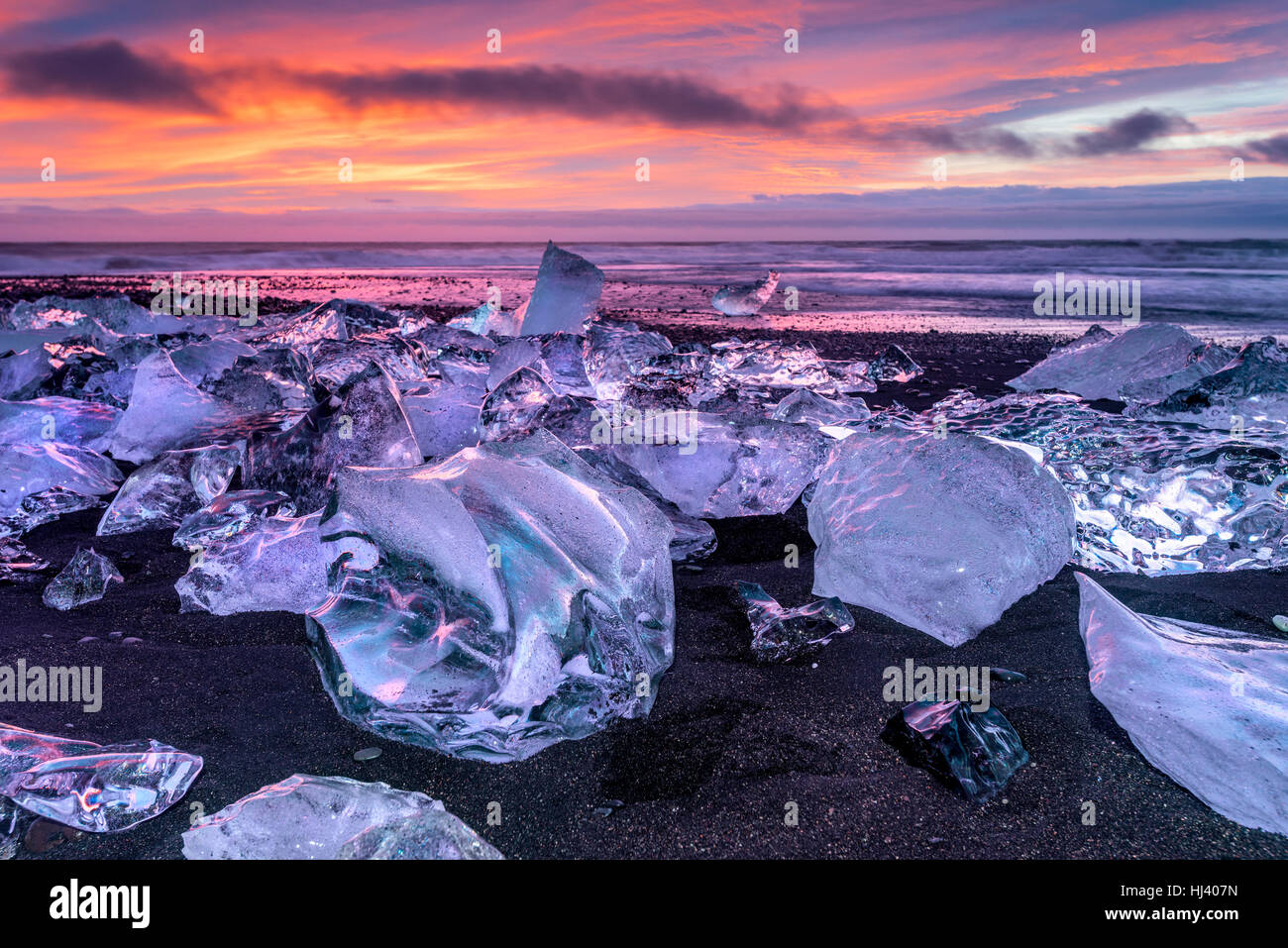 An iceberg along the shore of Jokulsarlon glacial lagoon during a vibrant red sunrise rests motionless as it is framed by cold ocean water. Stock Photo