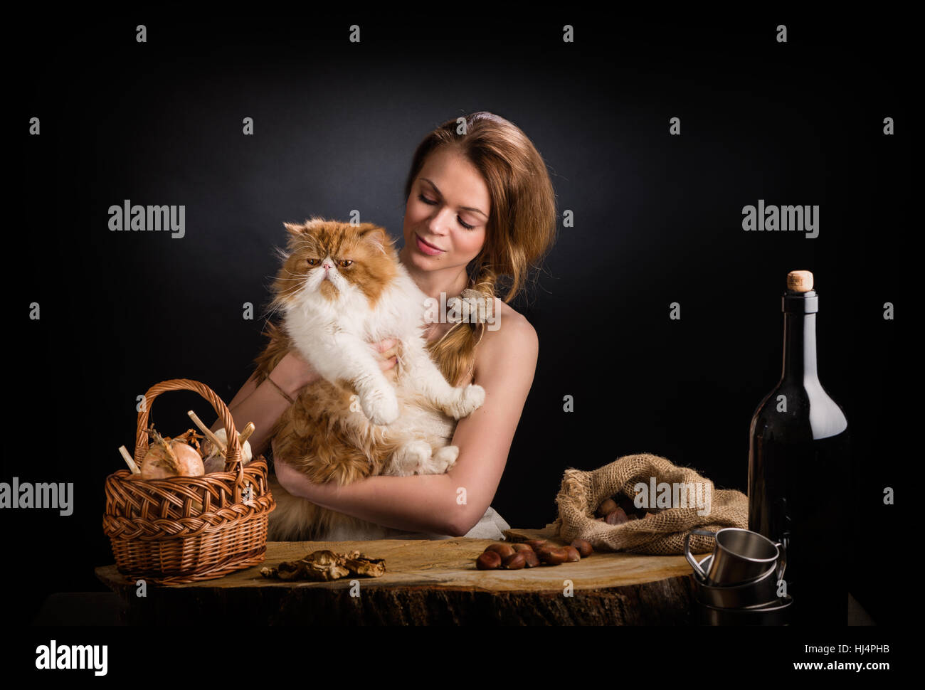 The young beautiful girl in a sundress from  canvas sits at an oak table with   remarkable red Persian cat on hands near  basket  onions , dried mushrooms and  bag  chestnuts Stock Photo