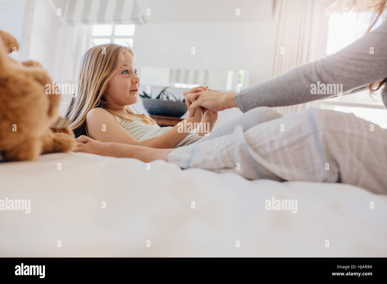 Shot of little girl lying on bed and laughing with her mother holding her hands. Girl playing with her mother in bedroom at home. Stock Photo