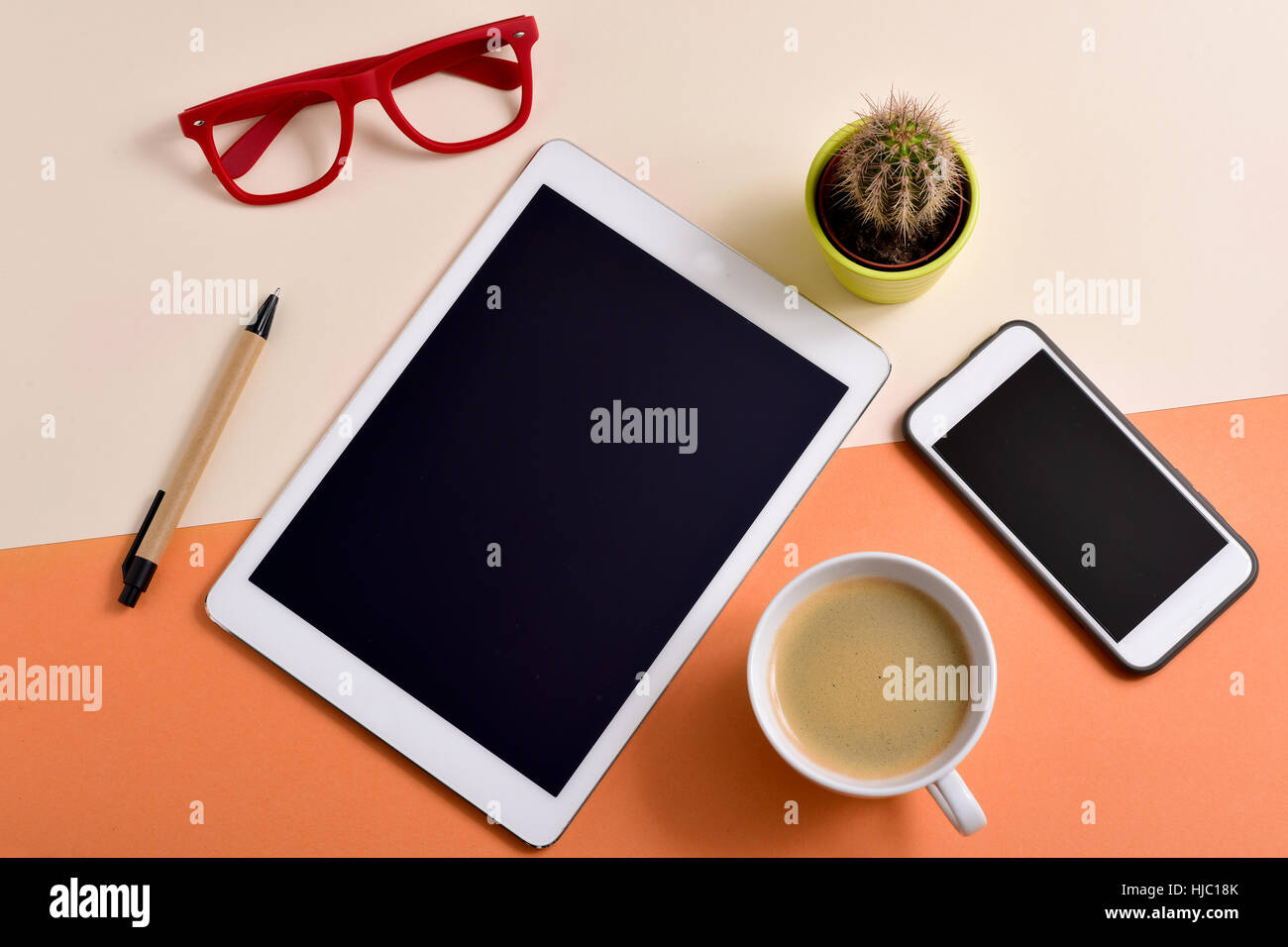 high-angle shot of an office desk full of things, such as a pair of red eyeglasses, a cup with white coffee, a tablet, a smartphone, a pen and a cactu Stock Photo